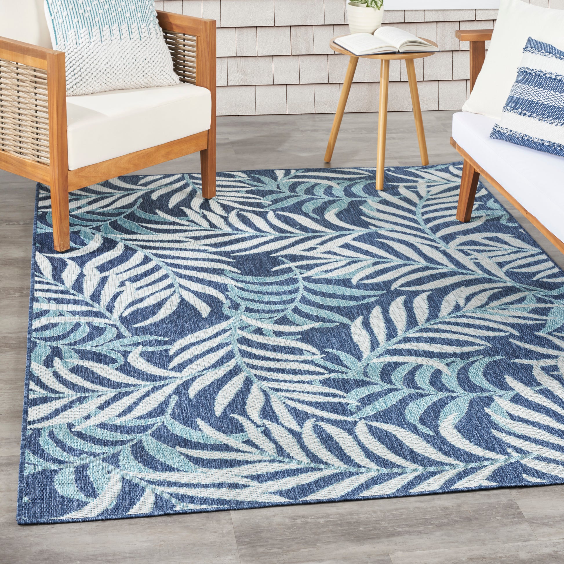 Rectangular blue tropical leaf-pattern rug with light blue and teal fronds on navy, woven texture, in a coastal living space with wood furniture.