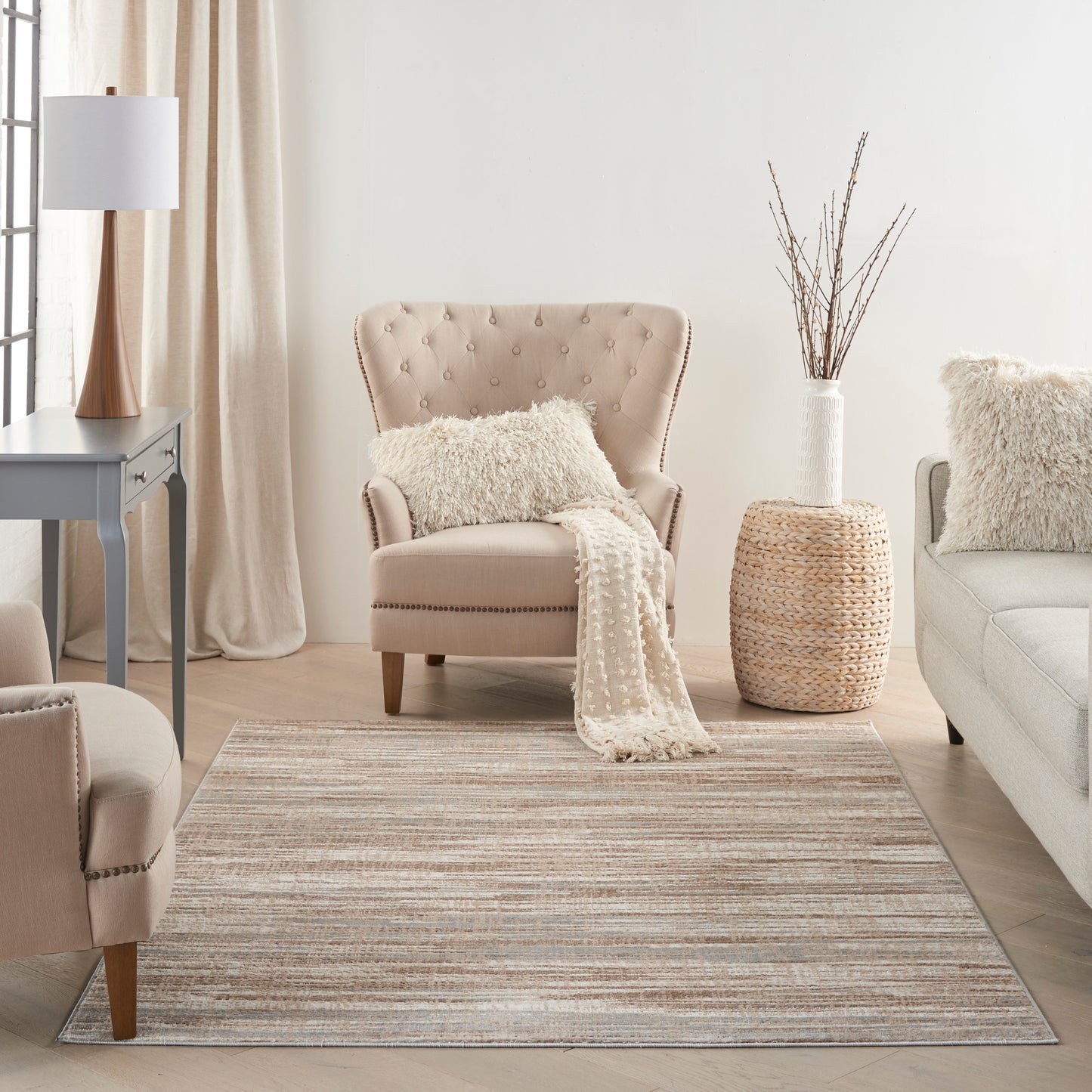 Beige tufted accent chair with cream knit blanket in a neutral living room; striped beige rug, woven side table, cream sofa.