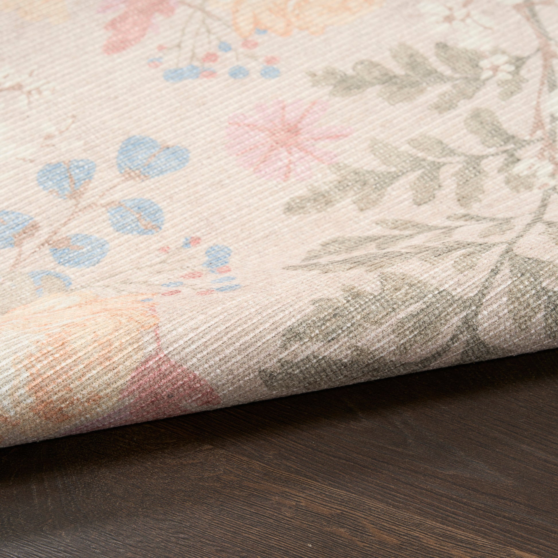 Pastel floral rug with pink, blue and peach blossoms and gray-green leaves on beige, folded over a dark wood surface.