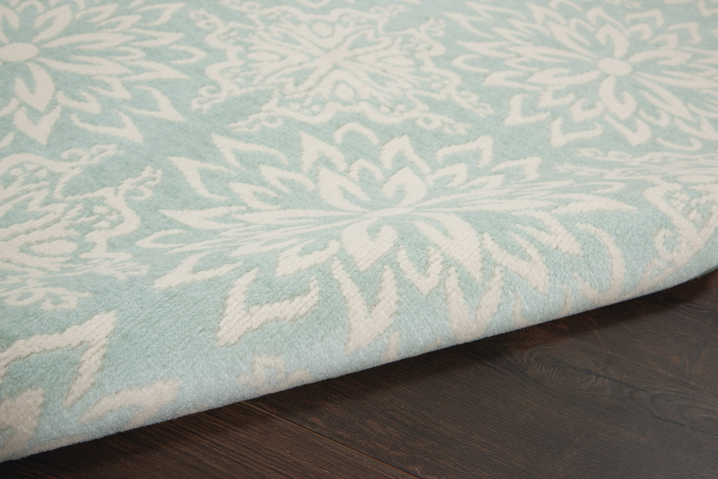 Light blue area rug with white floral pattern and thick pile, folded edge resting on dark wooden floor.