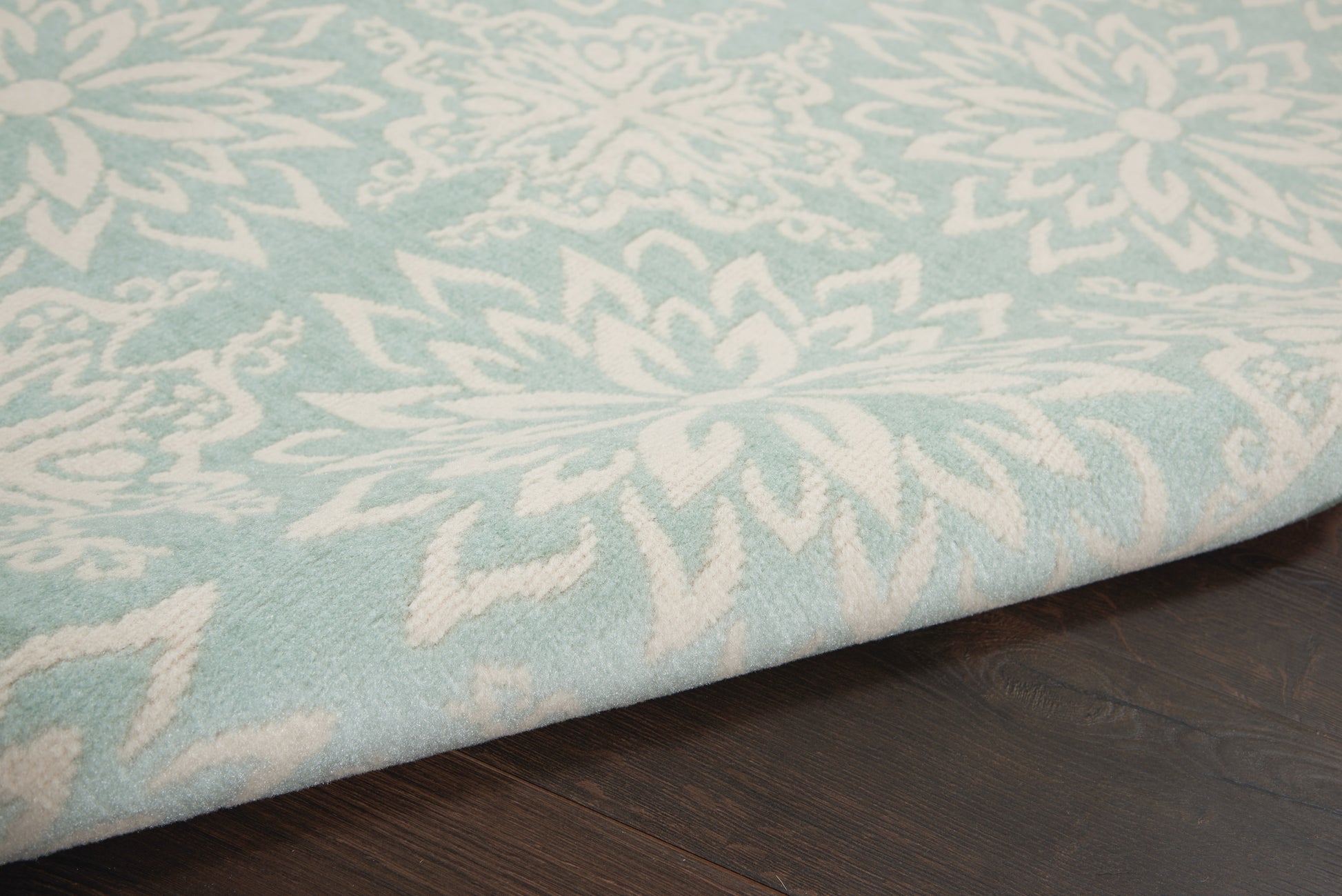 Light blue area rug with white floral pattern and thick pile, folded edge resting on dark wooden floor.
