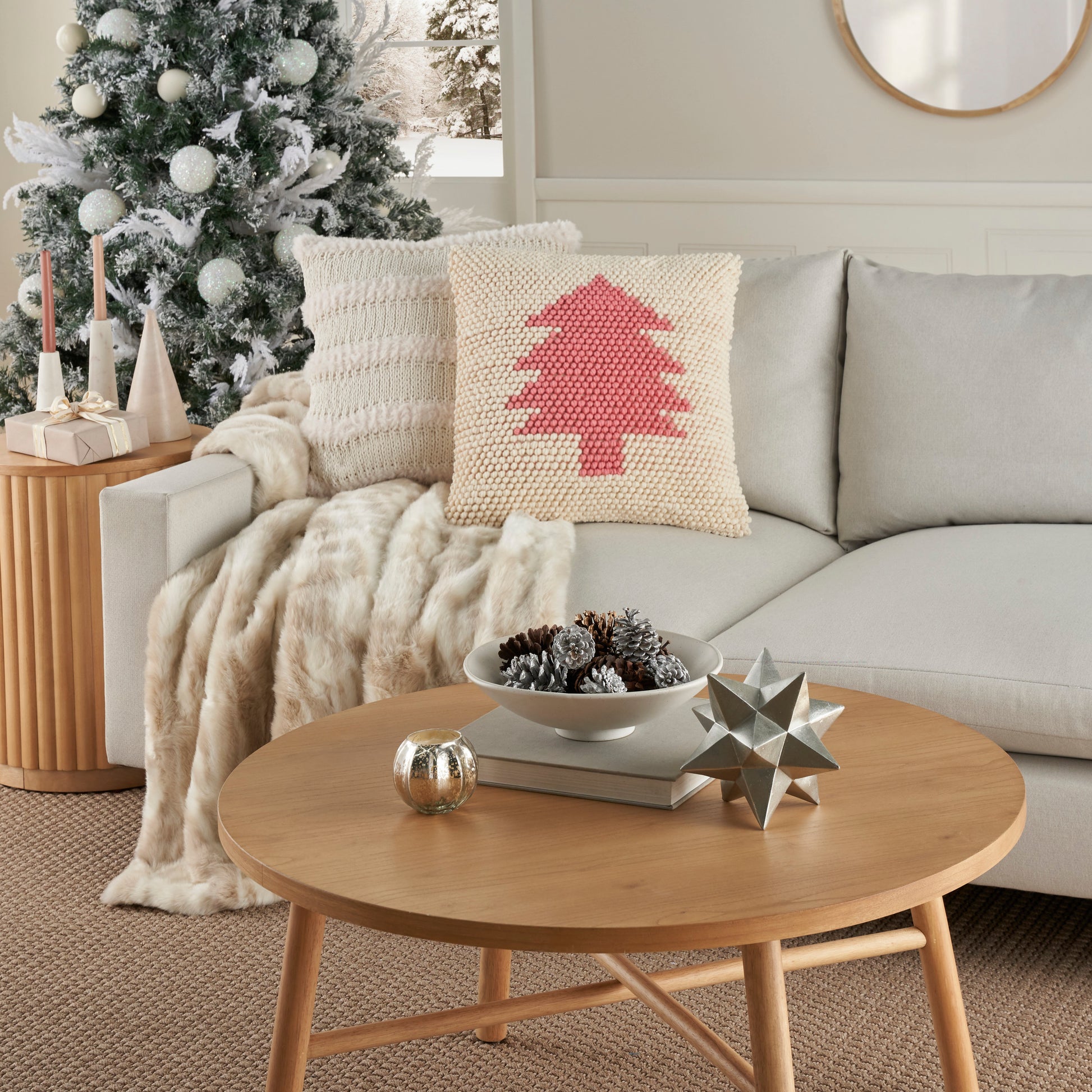 Round light-wood coffee table with star sculpture & pinecone bowl; beige sofa with knit pillow & cozy throw.