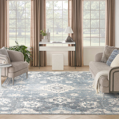 Cozy living room with blue patterned area rug, beige sofa, light curtains, white console table, lamp, & greenery.