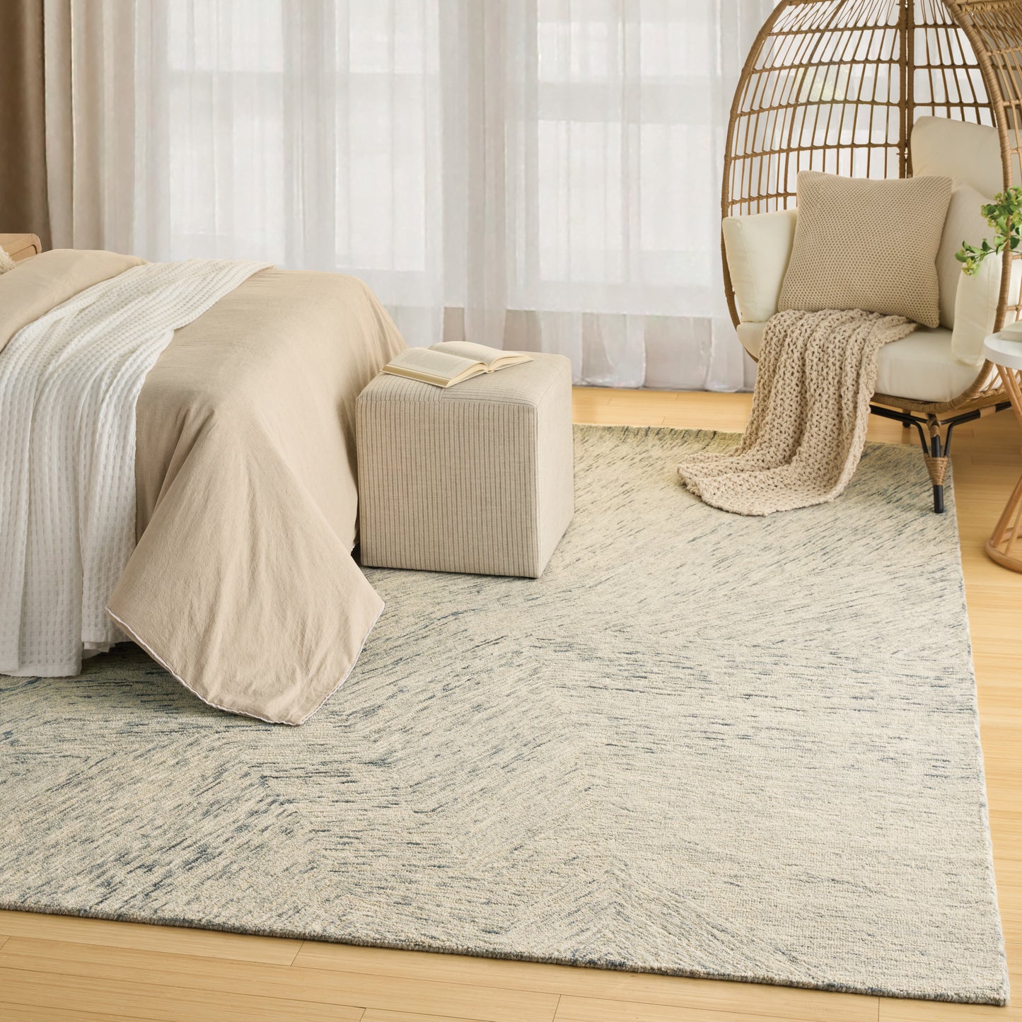 Neutral bedroom with beige bedding, woven rattan chair, soft throw, cream pouf, and a light patterned rug.