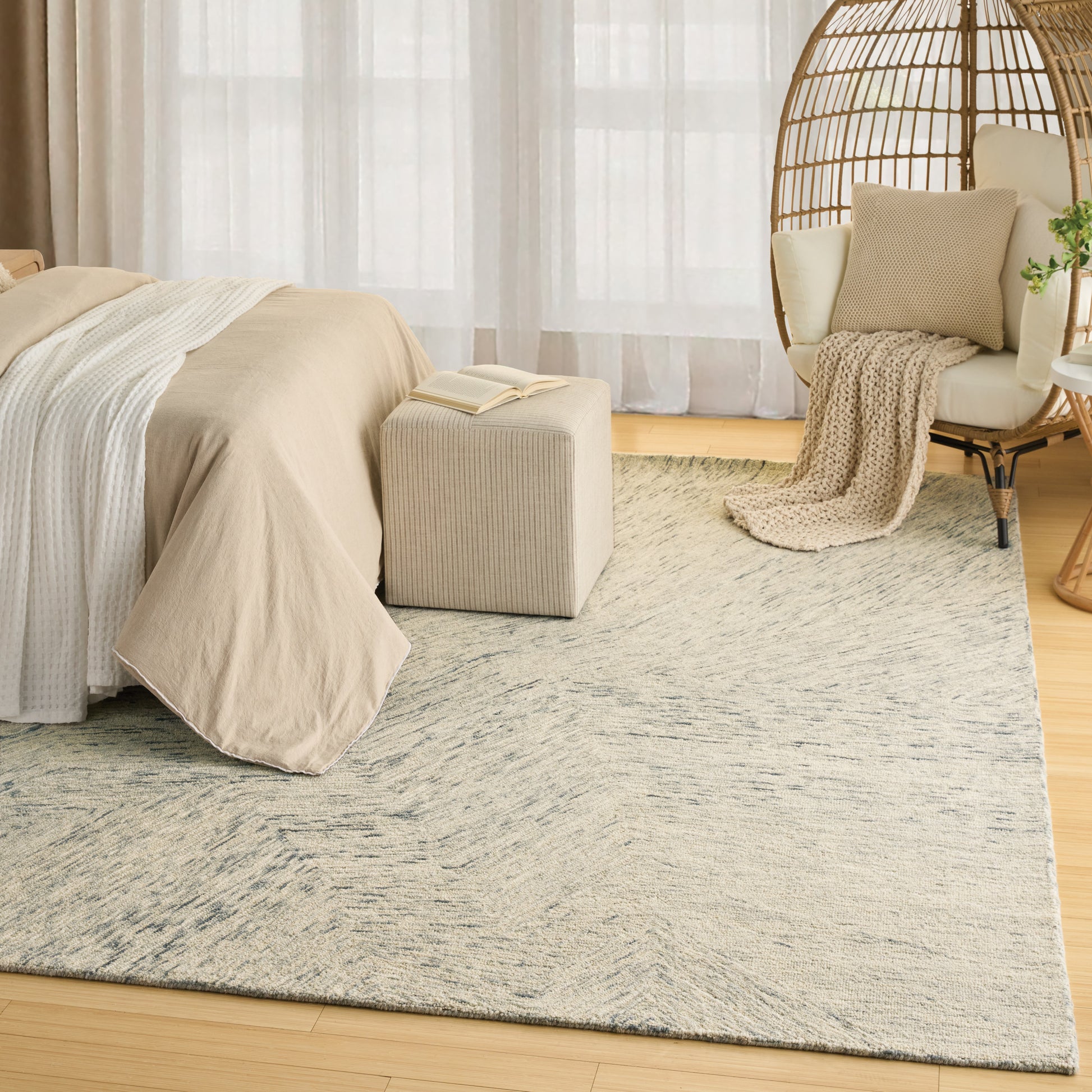 Neutral bedroom with beige bedding, woven rattan chair, soft throw, cream pouf, and a light patterned rug.