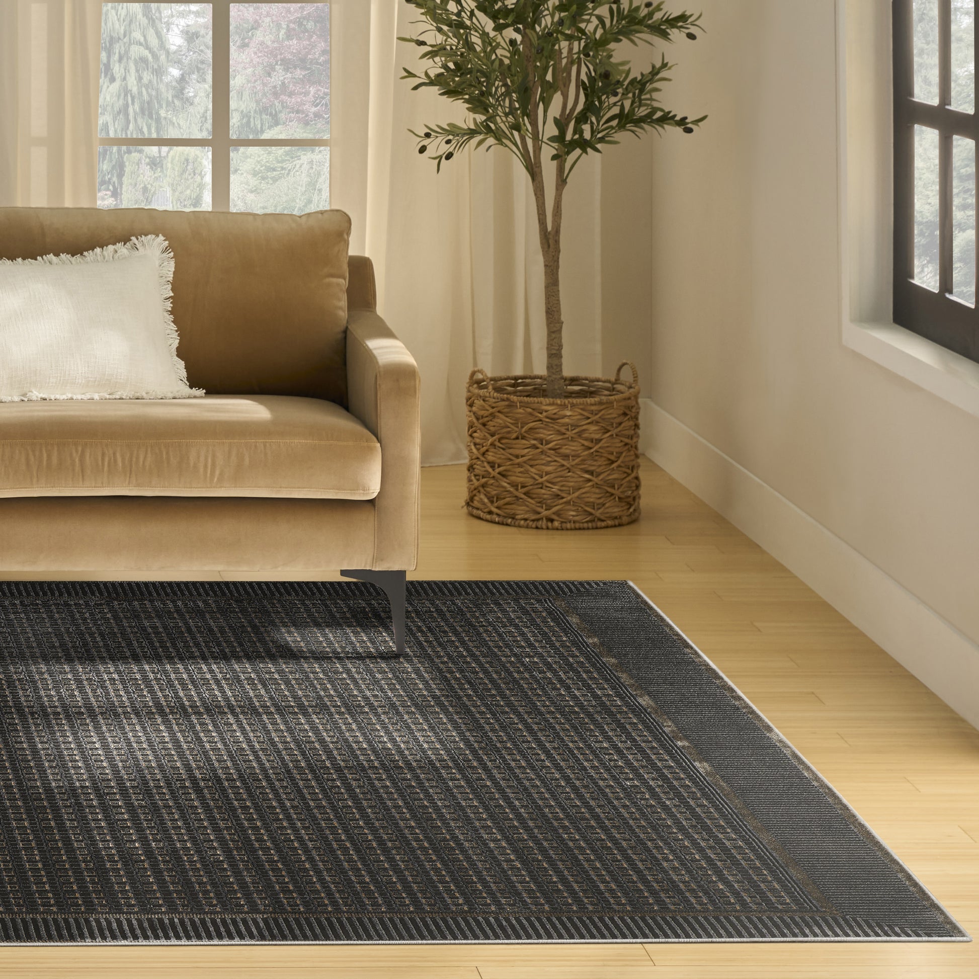 Beige sofa with white pillow, dark bordered geometric rug, and potted olive tree in a woven basket.