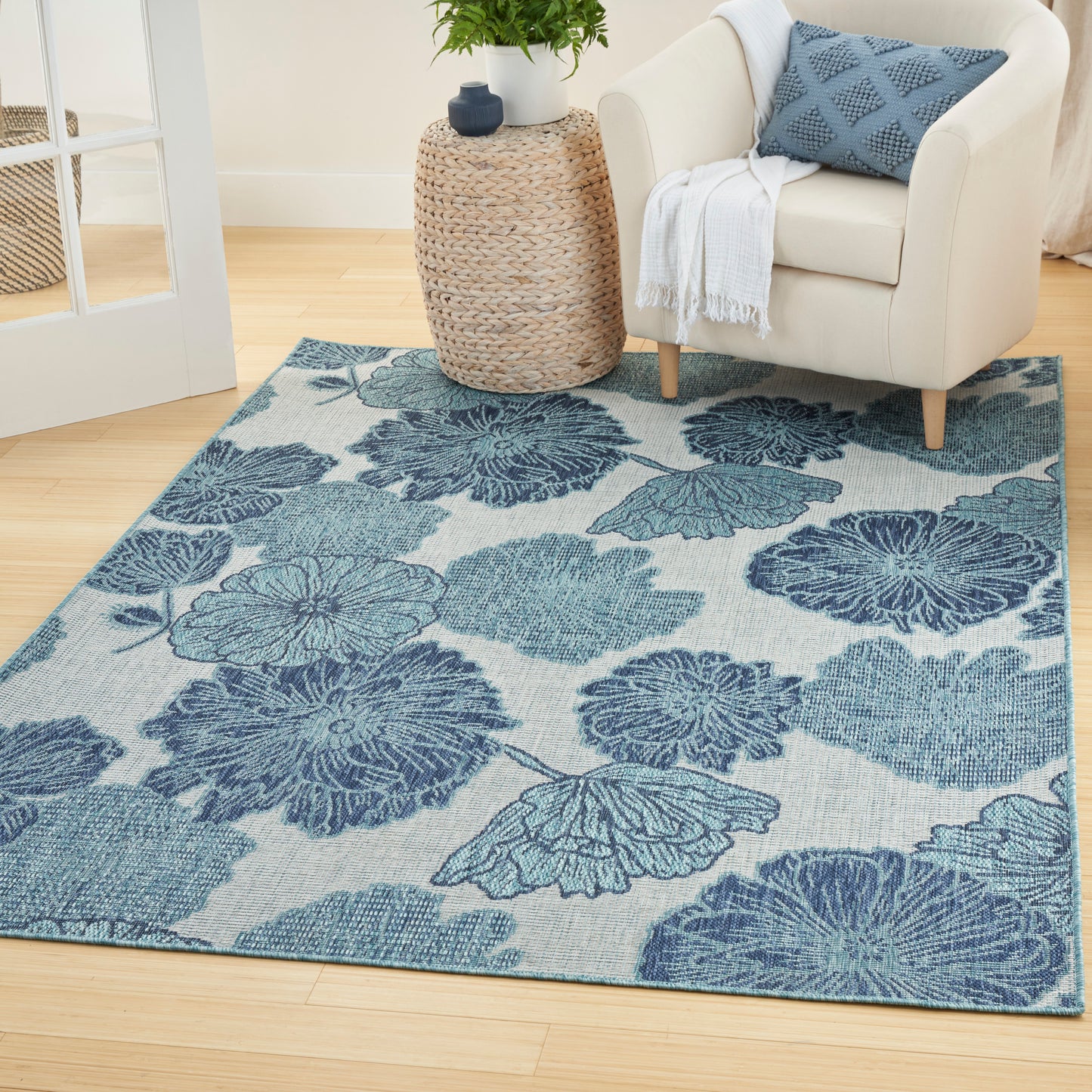 Blue floral area rug with large petal patterns on a beige backdrop; living room scene with a cream armchair and a wicker side table.
