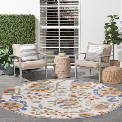 Outdoor seating area with beige cushioned metal chairs, round pastel floral rug, woven ottomans, and potted plants.