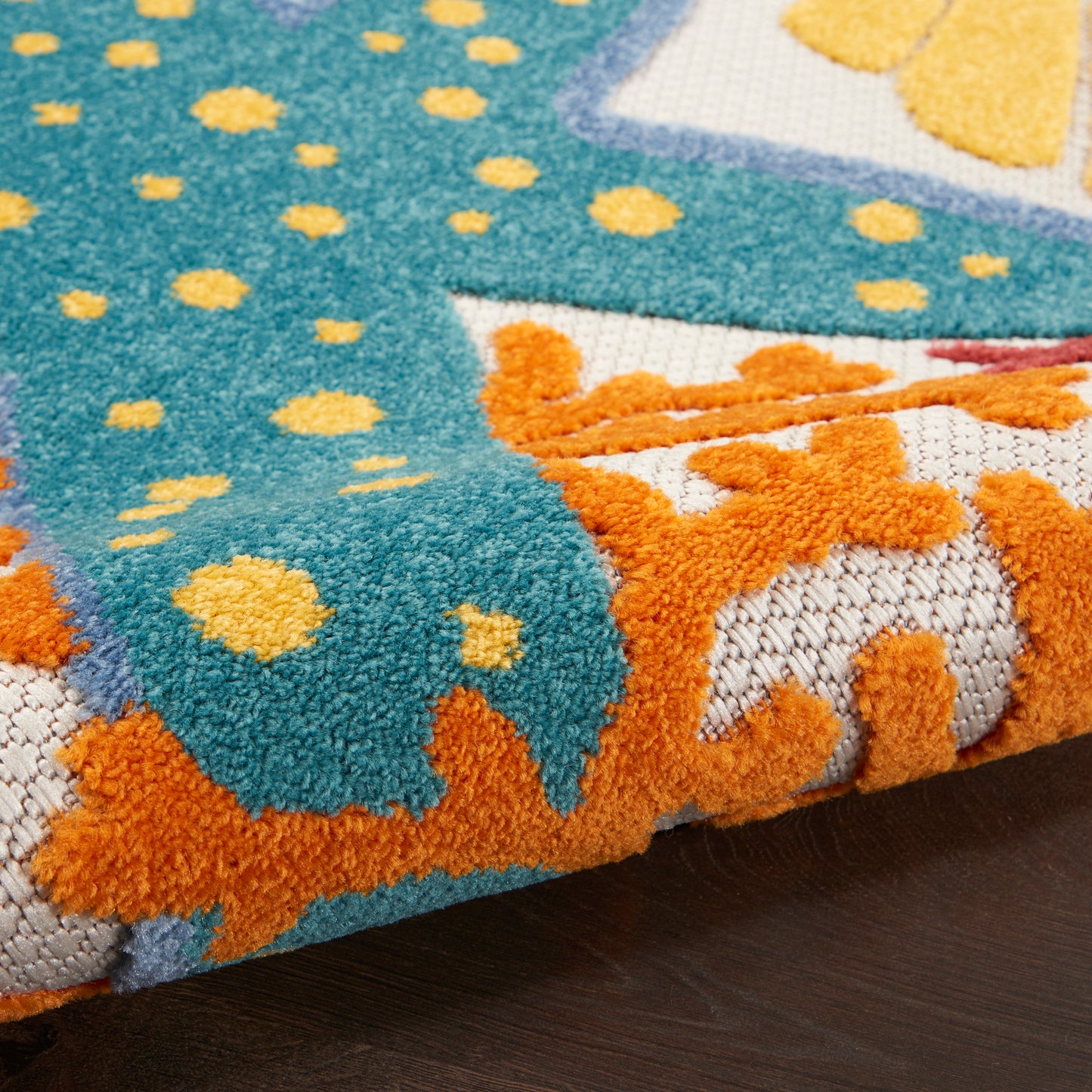 Close-up of turquoise tufted rug with yellow dots and orange border motif, folded edge on dark wooden floor.
