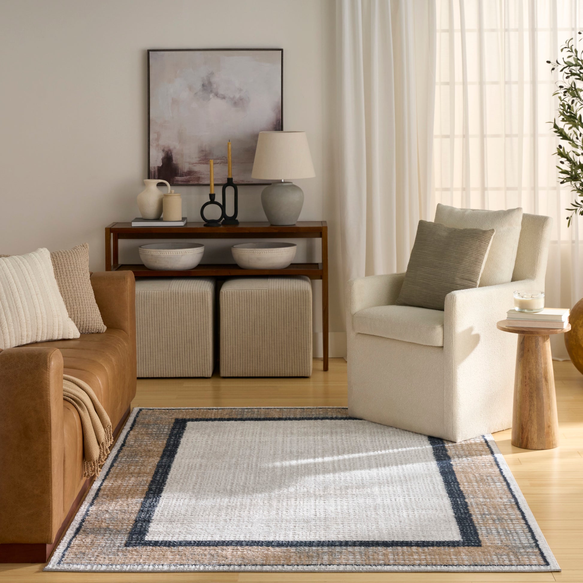 Neutral living room: tan leather sofa, cream armchair, beige rug with blue border, wooden console, lamp, and abstract art.
