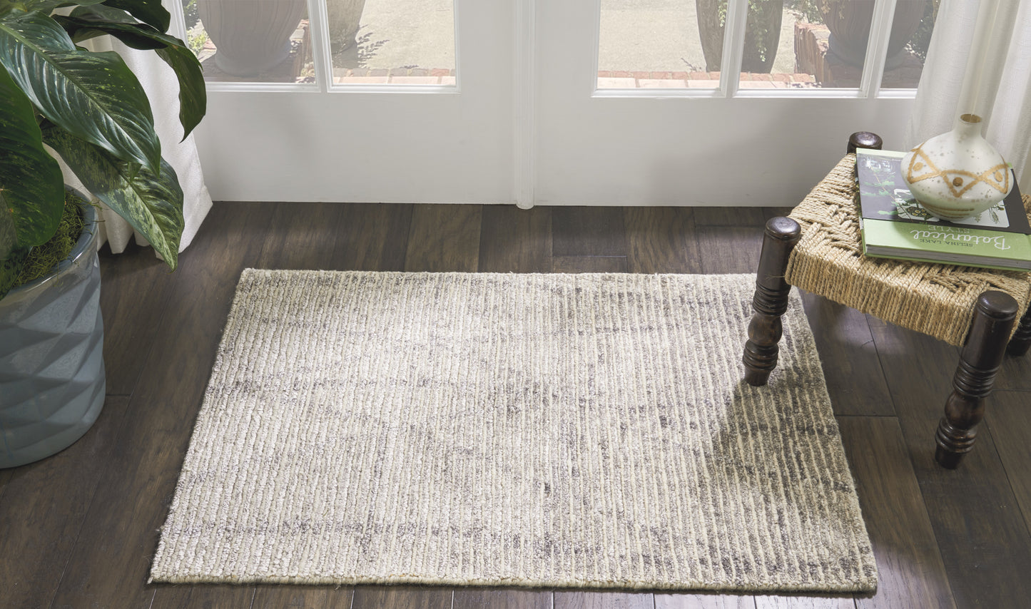 Light gray woven area rug on dark wood floor beside glass doors; potted plant left, wicker side table with books right.