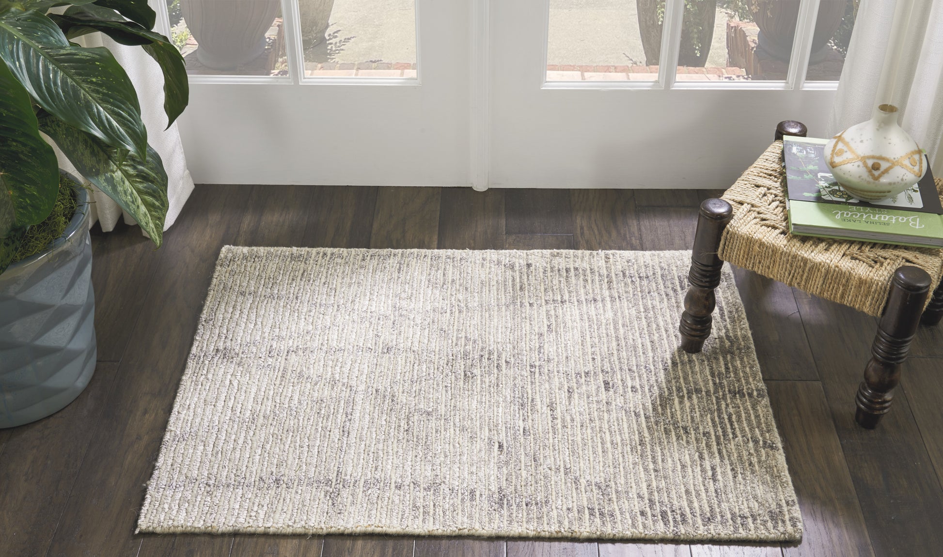 Light gray woven area rug on dark wood floor beside glass doors; potted plant left, wicker side table with books right.