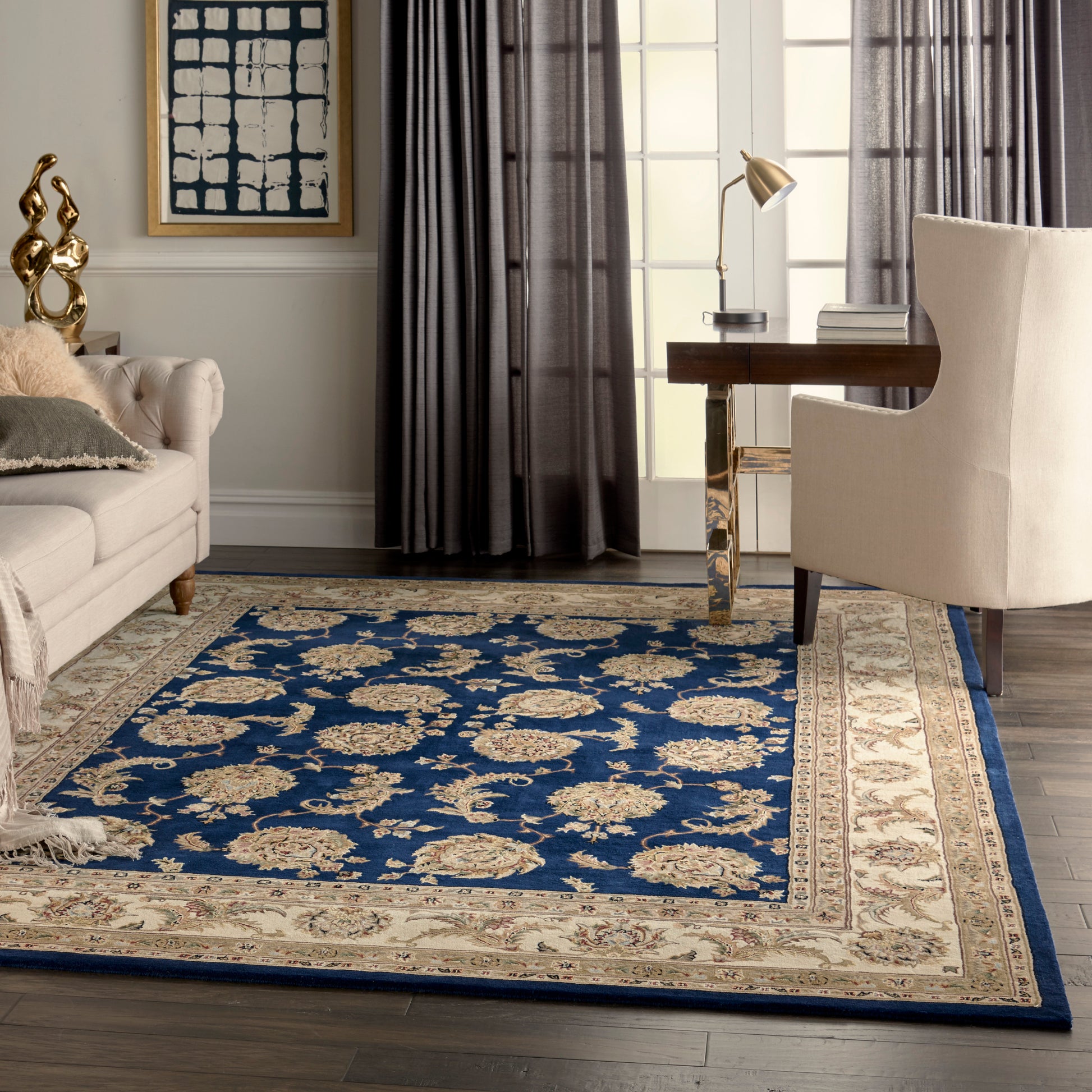 Blue ornate rug with beige floral pattern & border in a modern living room with cream sofa, beige armchair, & desk.