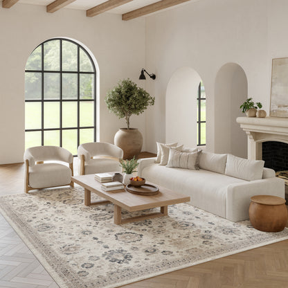 Neutral living room with cream sofa, curved armchairs, wooden coffee table, large arched window, patterned rug.