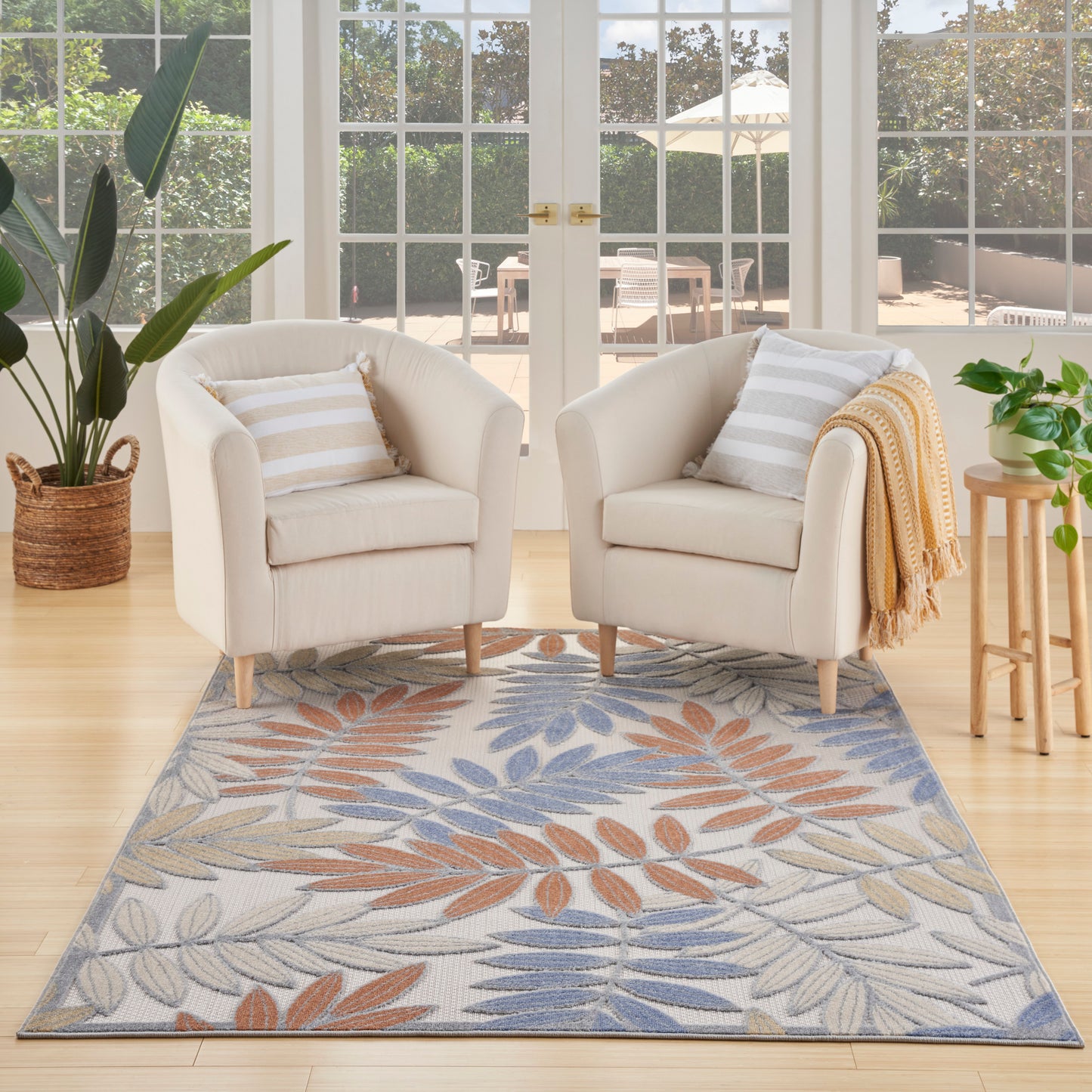 Outdoor rug 6' x 9' with blue-orange leaf pattern; two cream fabric armchairs with wooden legs, striped cushions, throw, and plants by glass doors.