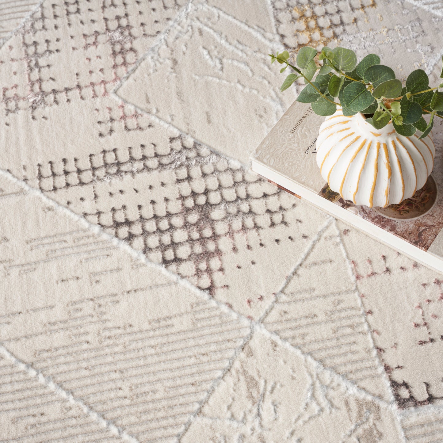 Beige patchwork woven rug with geometric motifs; ceramic ribbed vase with orange stripes and eucalyptus stems.