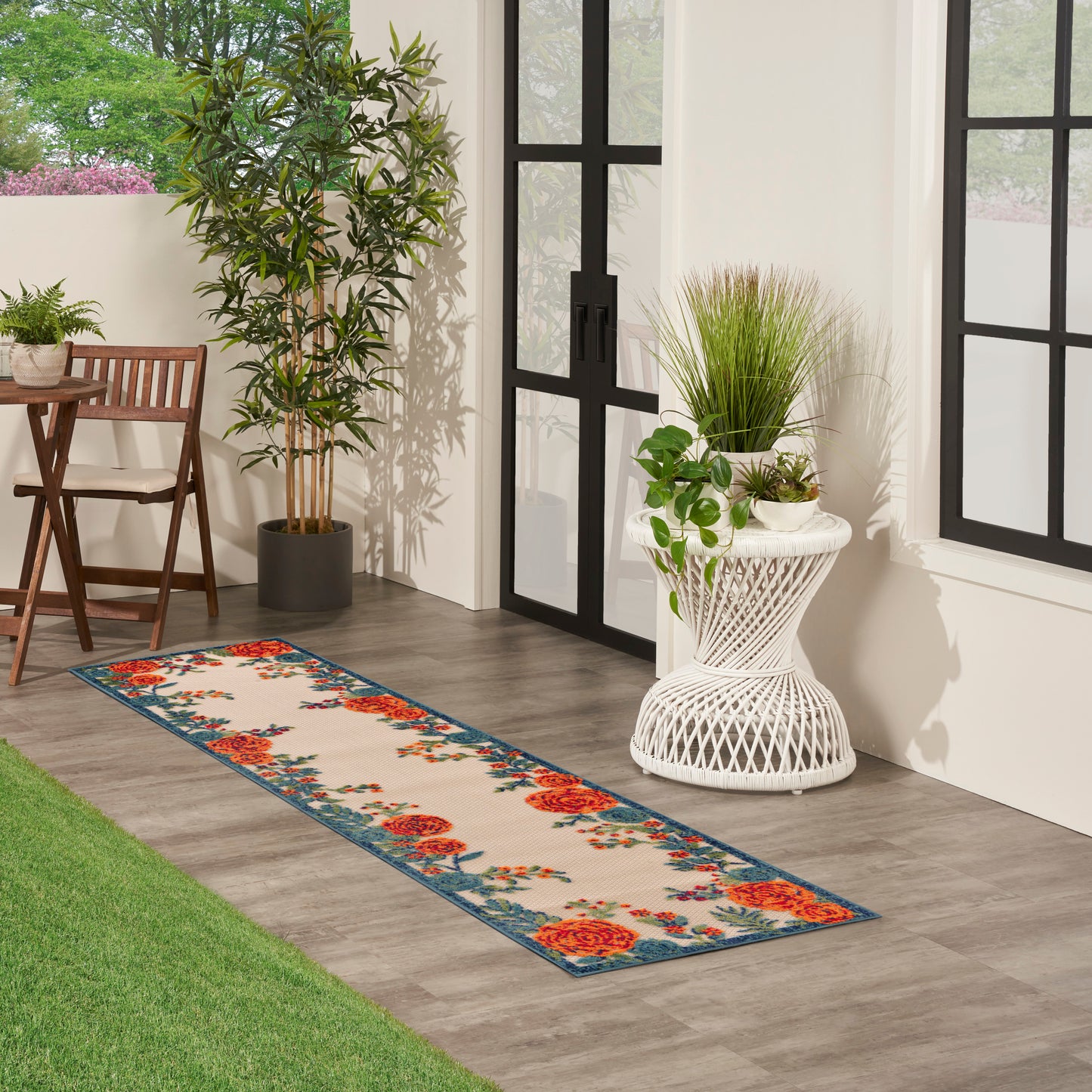 Floral runner rug with blue border on gray wood floor; outdoor porch with white wicker side table and potted plants.