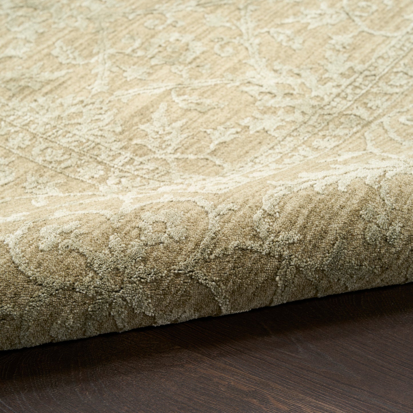 Beige damask patterned area rug with plush pile and folded edge, resting on dark wood floor.