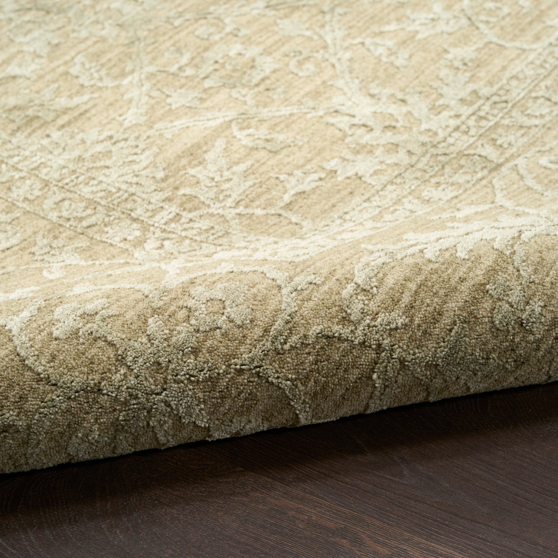 Beige damask patterned area rug with plush pile and folded edge, resting on dark wood floor.