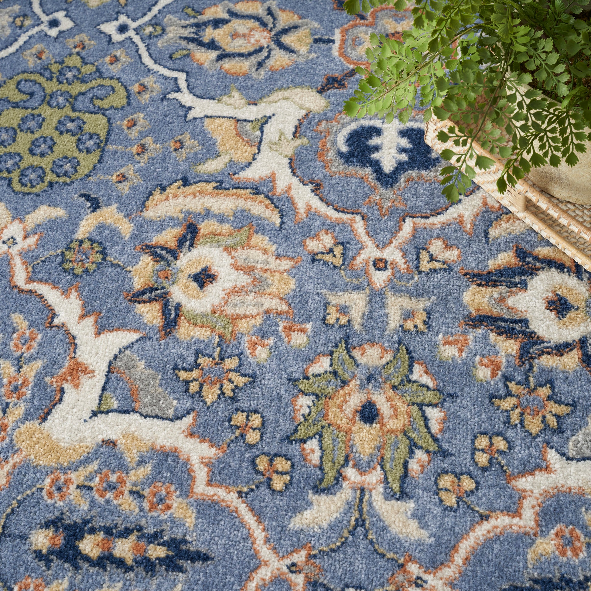 Blue floral-pattern rug with cream, orange & green motifs; plush textured field with a plant in a wicker basket in the corner.