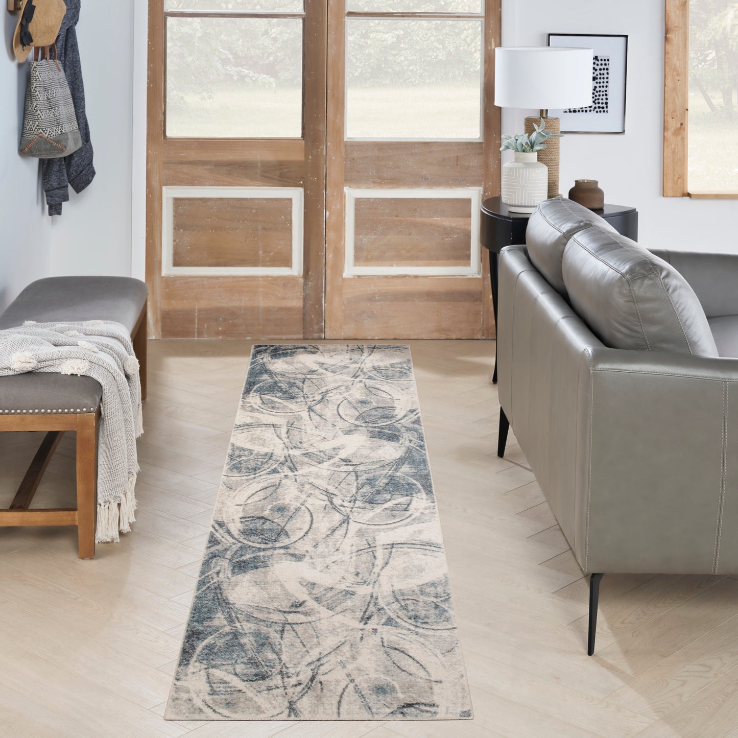 Entryway with blue-gray abstract patterned rug runner, gray leather sofa, wooden doors, bench with throw blanket.