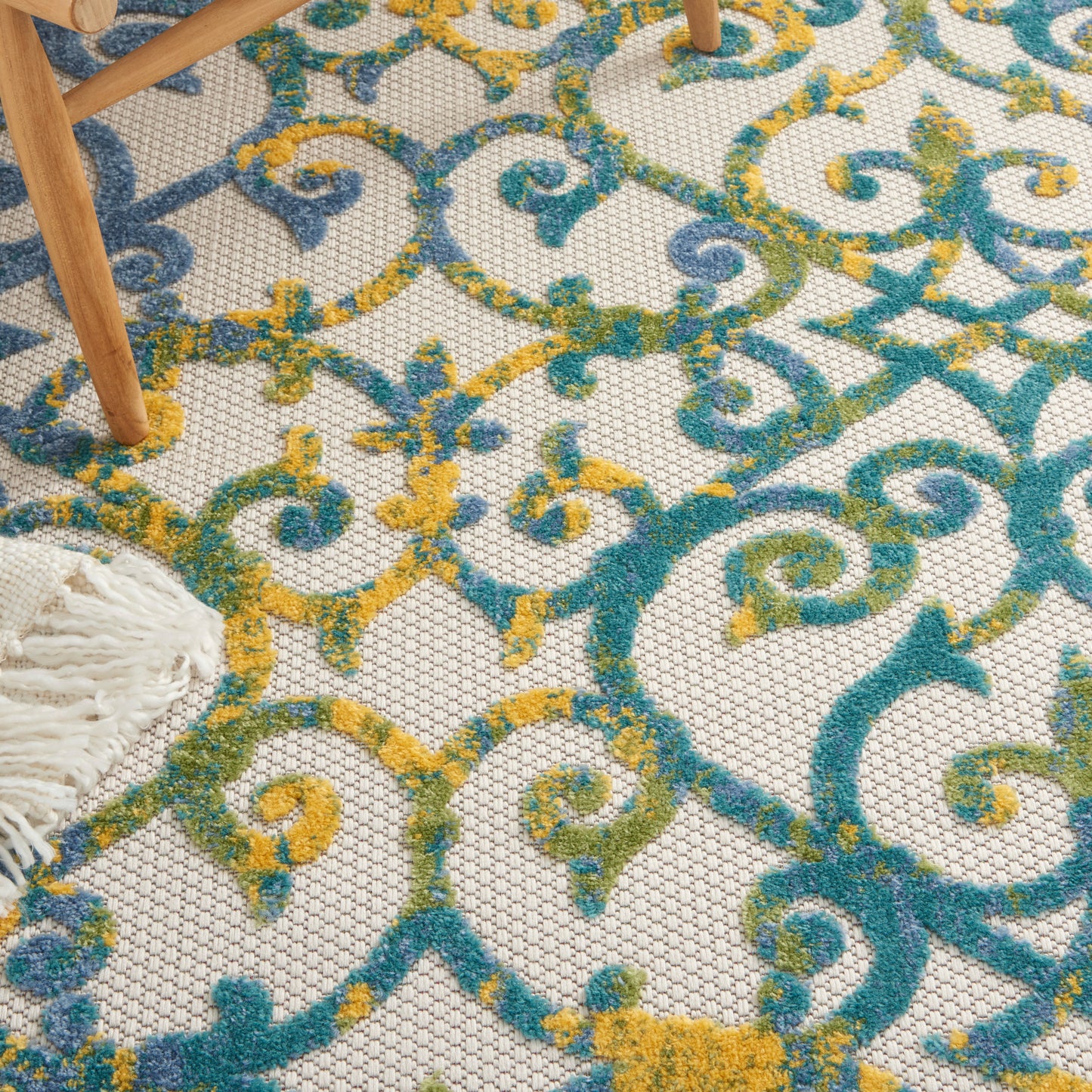 Colorful ornate rug with teal, blue, and mustard scrollwork on cream lattice; wooden chair legs and white fringe visible.