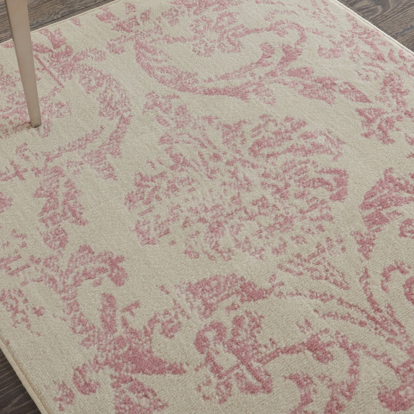 Beige rug with pink damask floral pattern; soft texture with wooden floor edge and table leg visible.