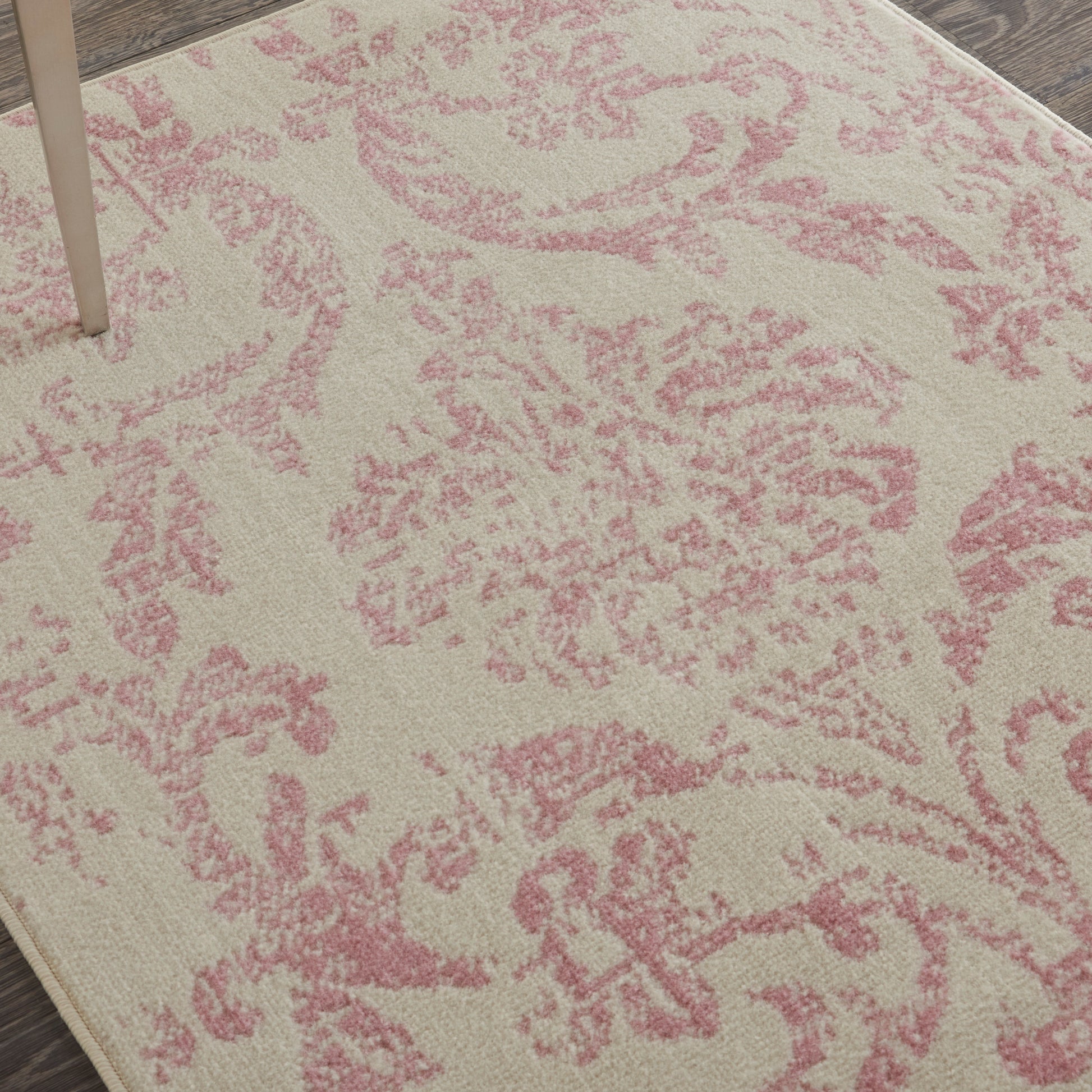 Beige rug with pink damask floral pattern; soft texture with wooden floor edge and table leg visible.