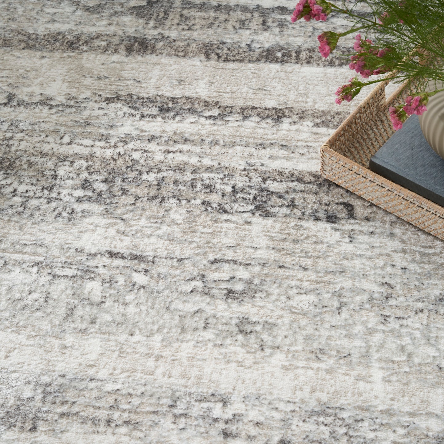 Beige-gray textured area rug with a woven basket containing a dark notebook and pink flowers in the corner.