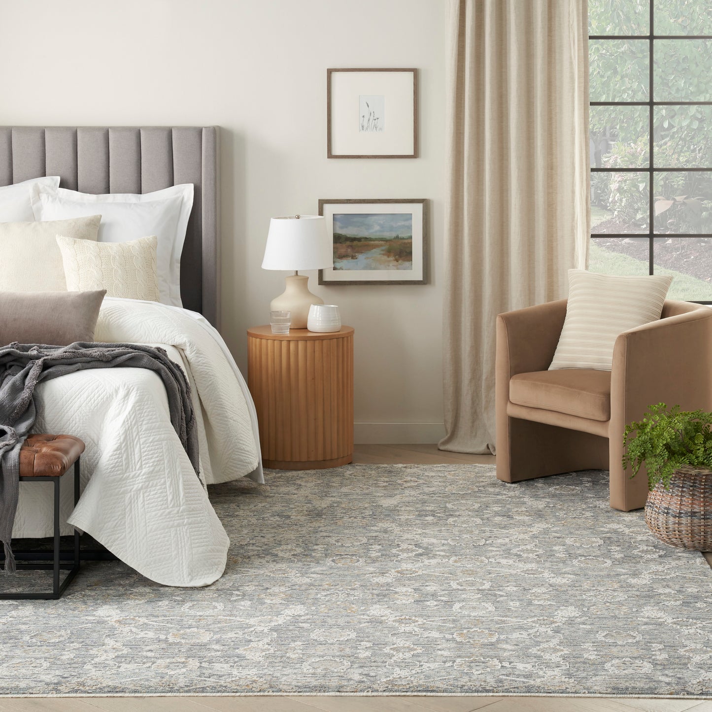 Neutral bedroom with gray upholstered headboard, white bedding, beige armchair, round wood nightstand, lamp, rug.