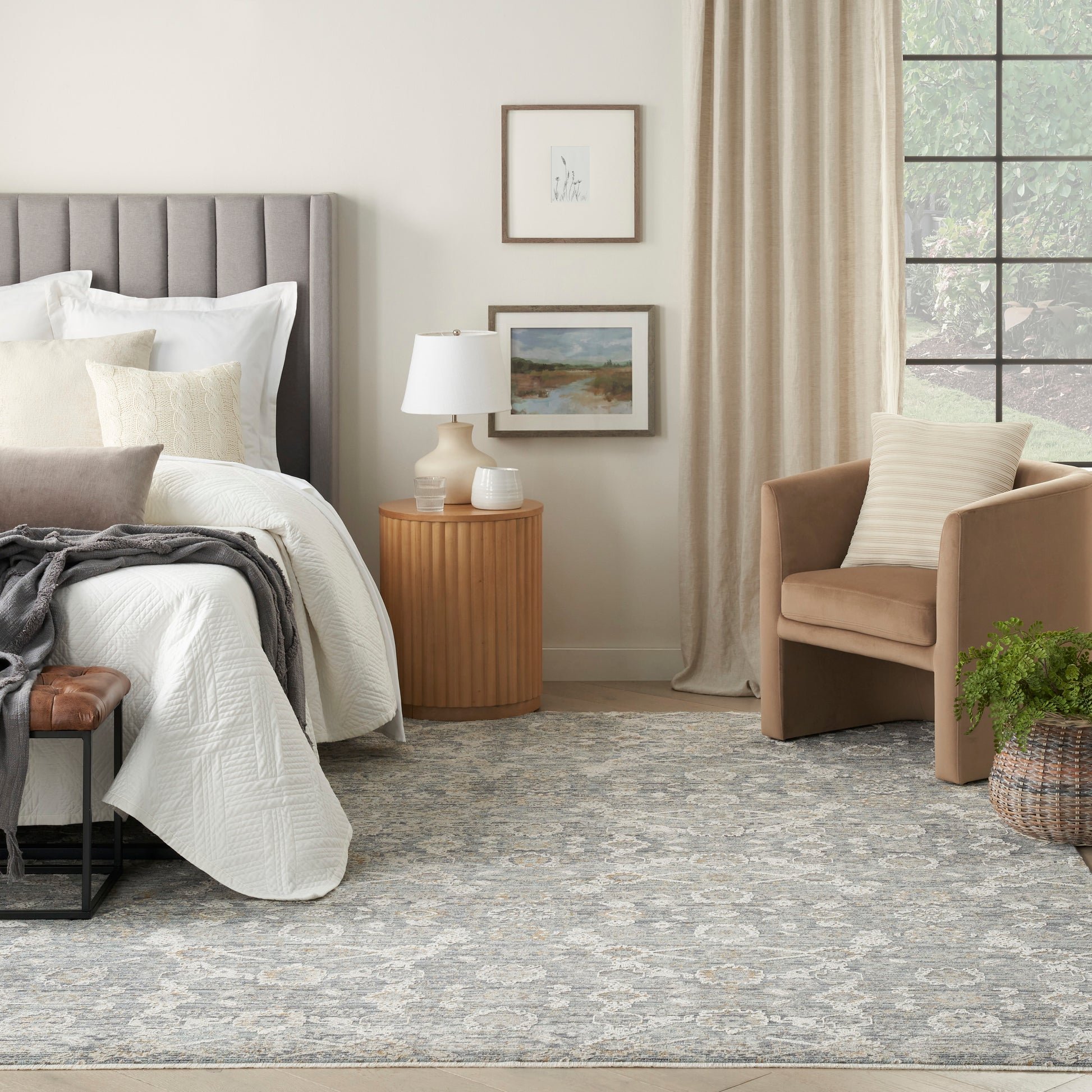 Neutral bedroom with gray upholstered headboard, white bedding, beige armchair, round wood nightstand, lamp, rug.