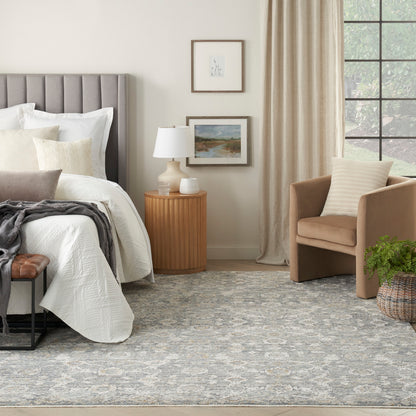 Neutral bedroom with gray upholstered headboard, white bedding, beige armchair, round wood nightstand, lamp, rug.