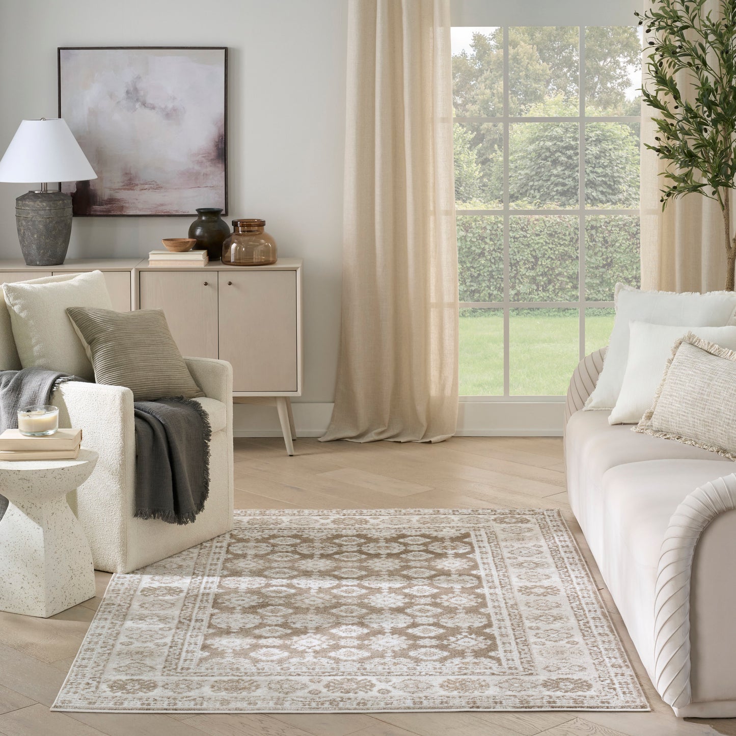 Neutral living room with beige sofa, cream chair, patterned rug, light wood sideboard, abstract art, and sheer curtains.