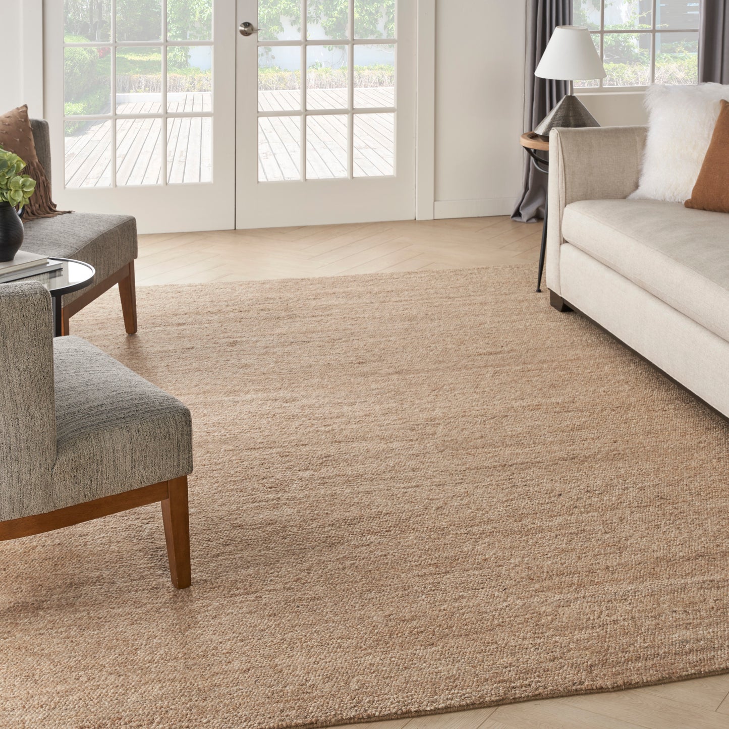 Natural jute area rug anchors a bright living room with gray sofa, beige chair, round glass table, and French doors