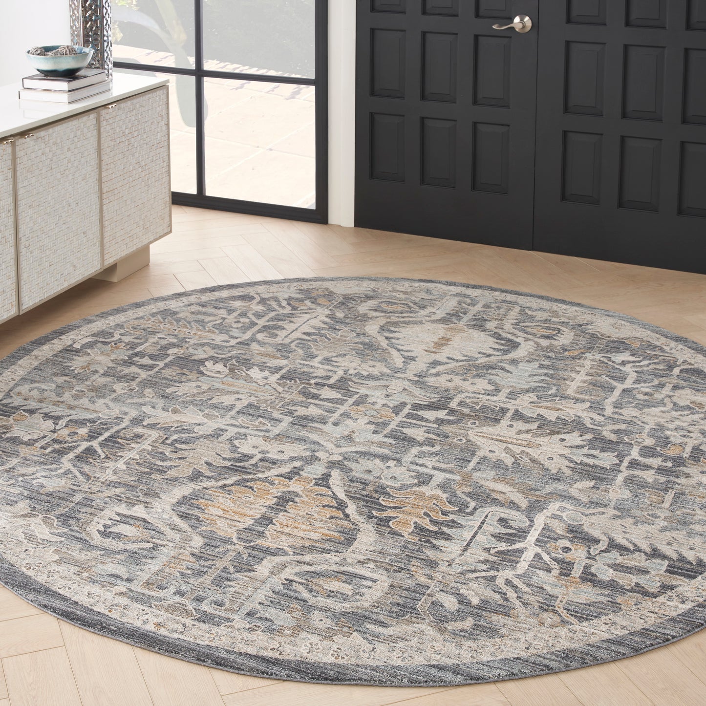 Round blue-gray woven rug with beige pattern, traditional oriental style, on light wood floor beside dark paneled doors.