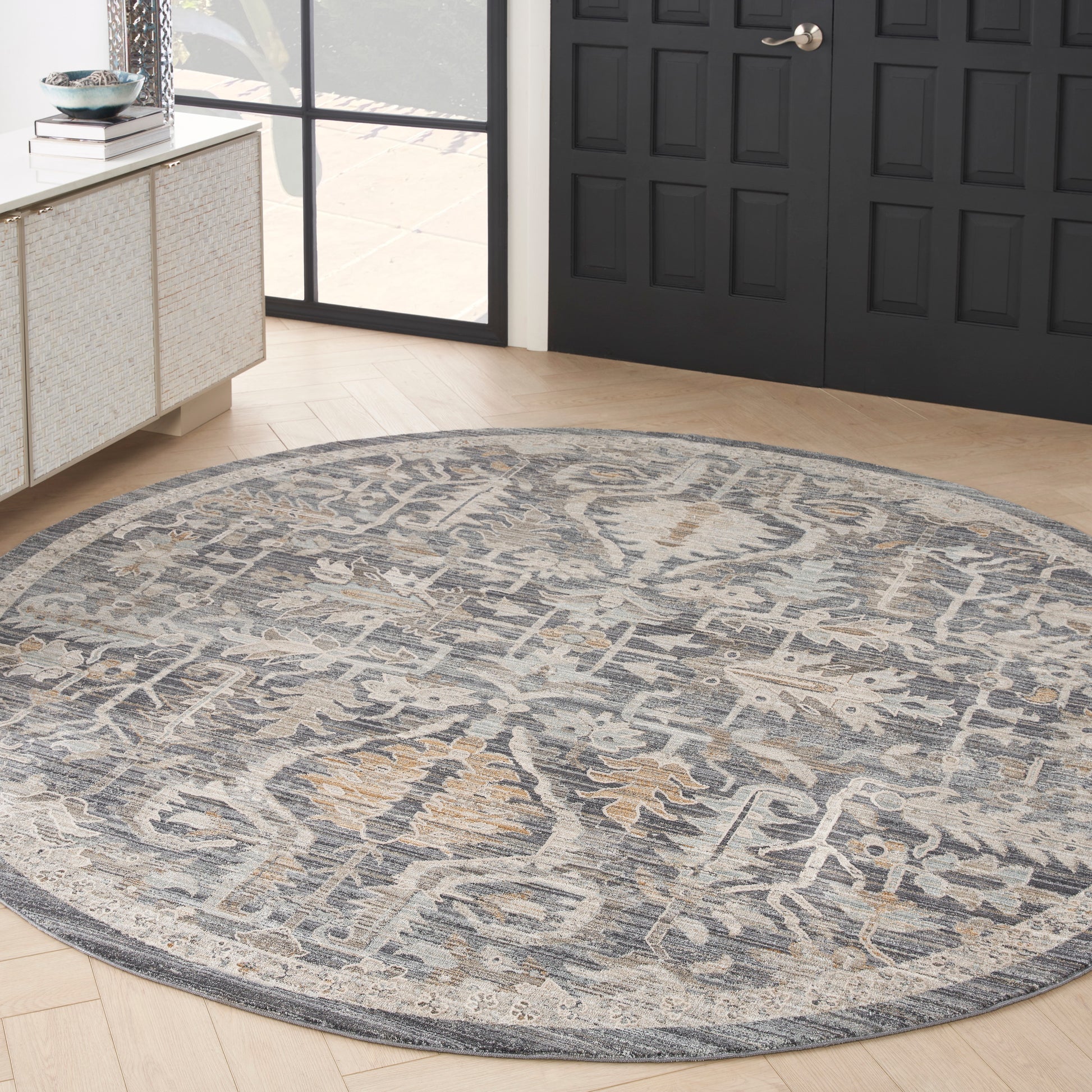 Round blue-gray woven rug with beige pattern, traditional oriental style, on light wood floor beside dark paneled doors.