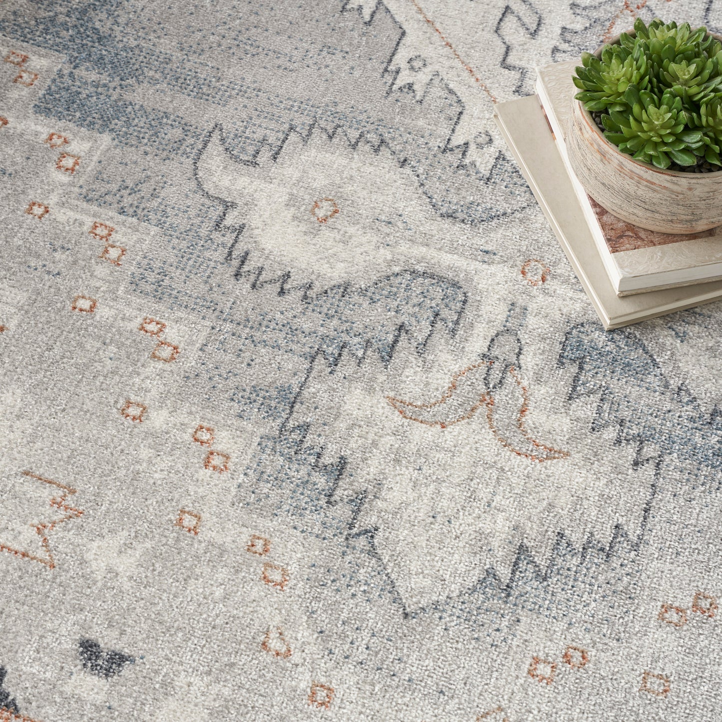 Soft gray-blue rug with abstract geometric pattern in blue and orange, shown with a succulent on books.