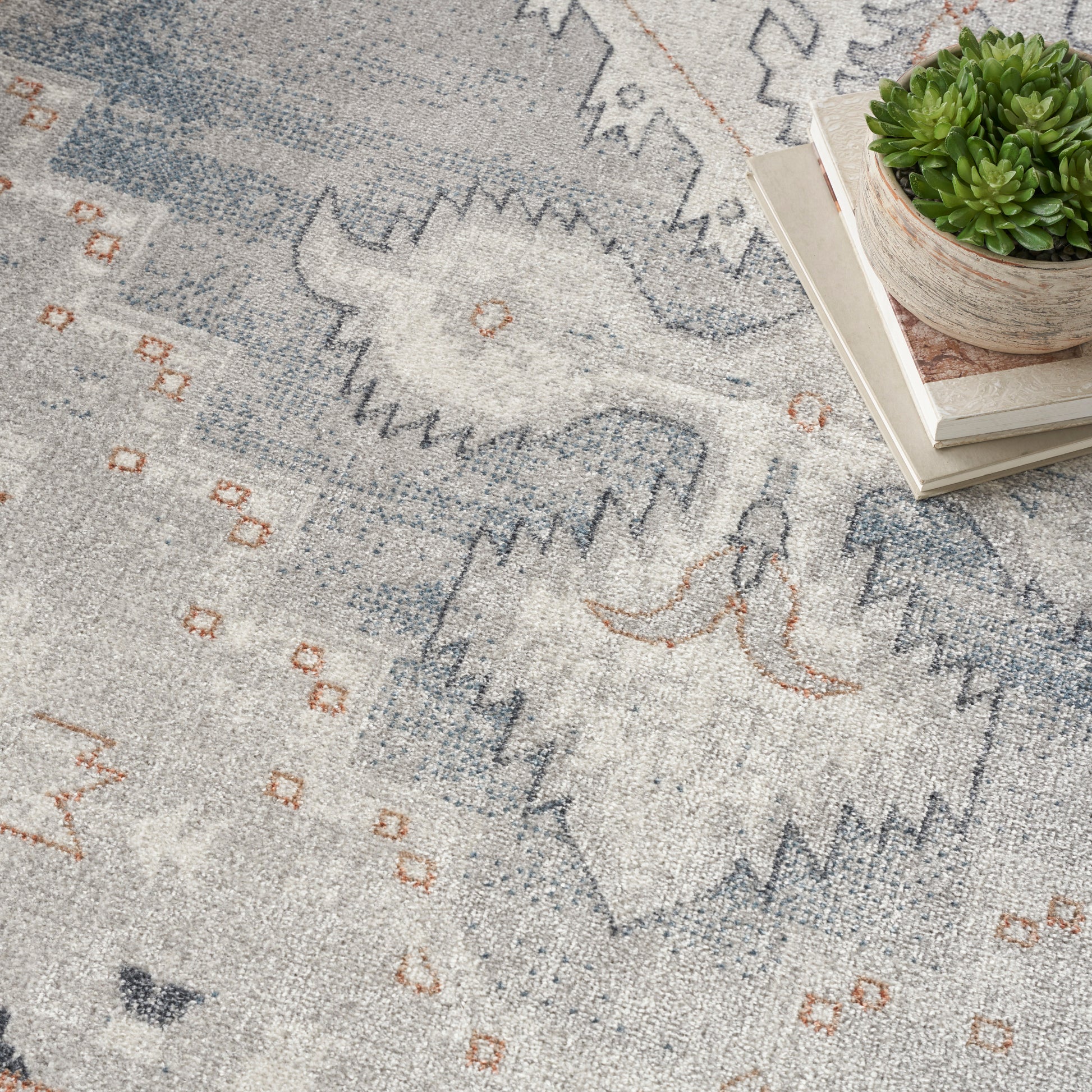 Soft gray-blue rug with abstract geometric pattern in blue and orange, shown with a succulent on books.