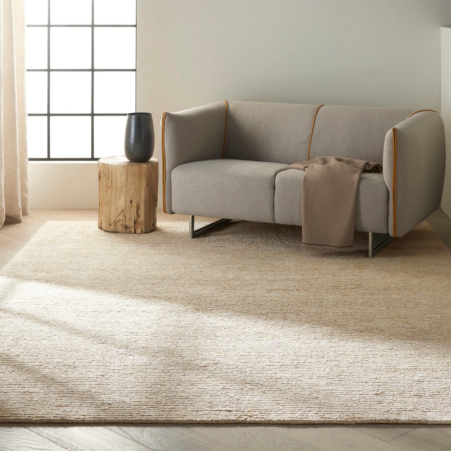Light natural fiber rug with subtle texture; a gray sofa with orange piping, beige throw, wooden stump table and a black vase sit on it.