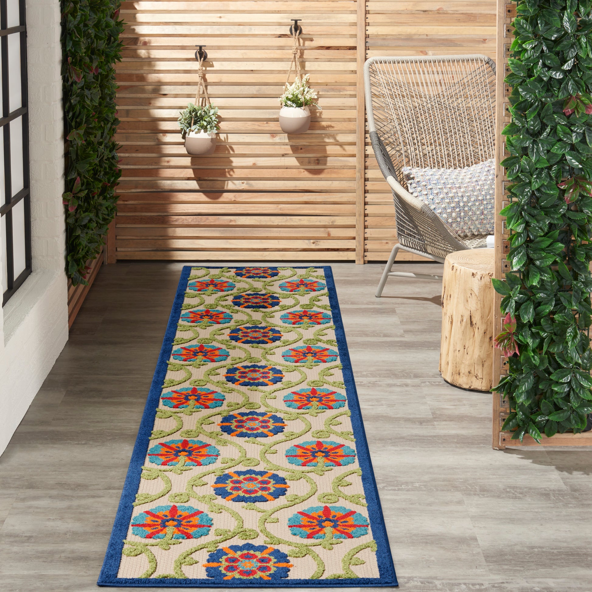 Narrow outdoor porch with long beige floral runner rug with blue border, wicker chair, hanging planters, and wood slat wall.