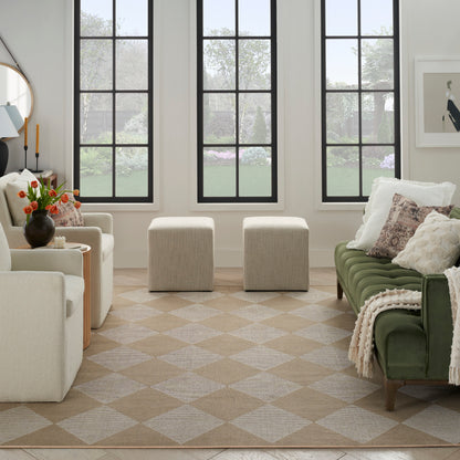 Neutral-toned living room with green velvet sofa, beige armchairs, cream ottomans, diamond rug, and vase of orange flowers.