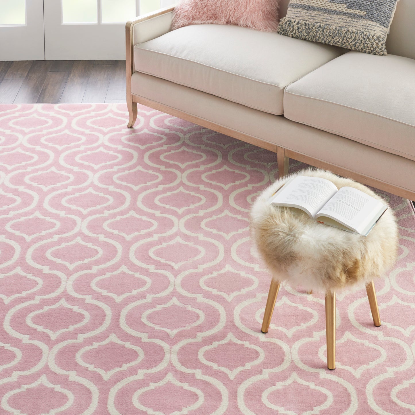 Pink geometric trellis rug, fluffy sheepskin stool with wood legs, beige sofa, pastel cushions, cozy living room.