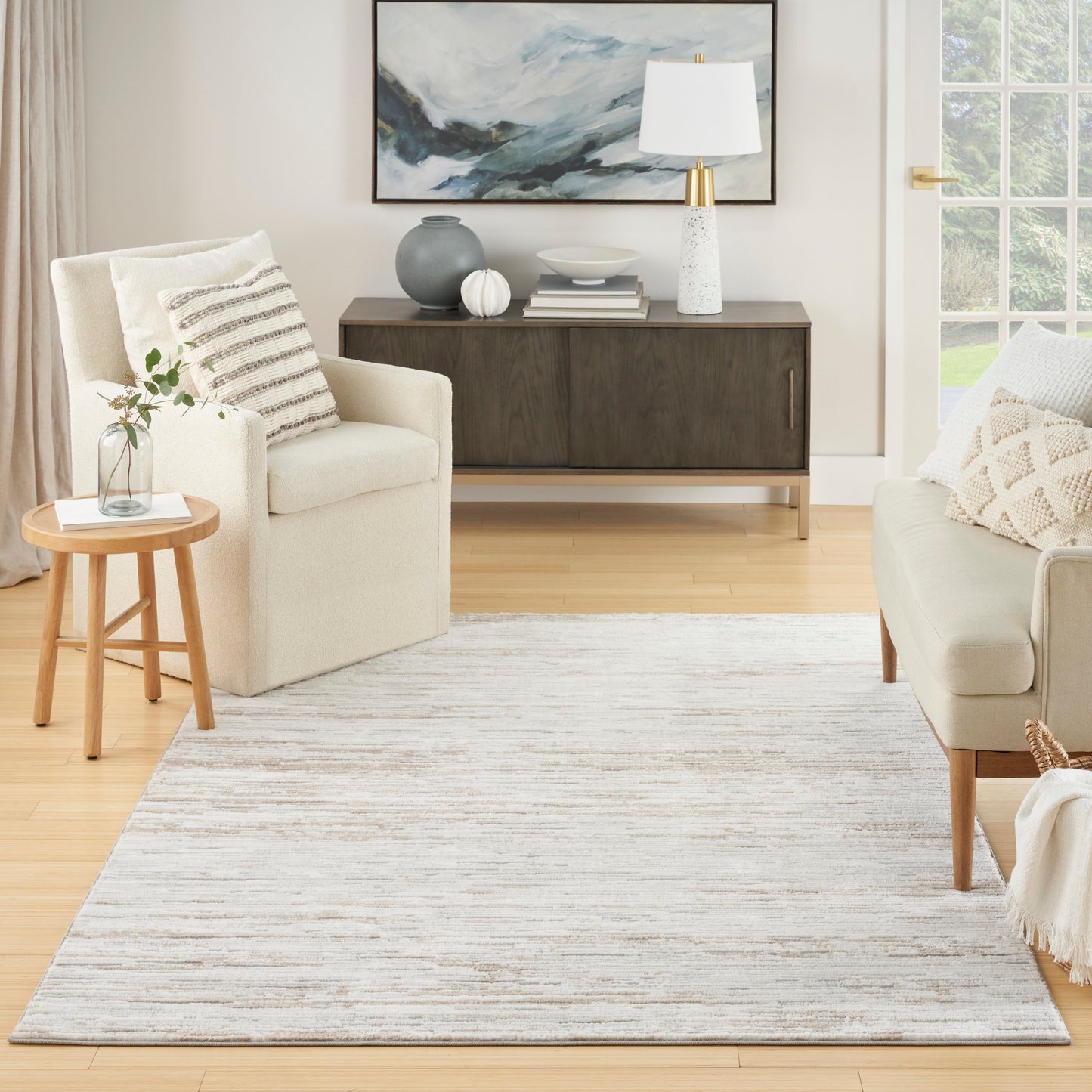 Neutral living room with cream armchairs, light gray rug, round wood side table, dark wood console, abstract art, white lamp.