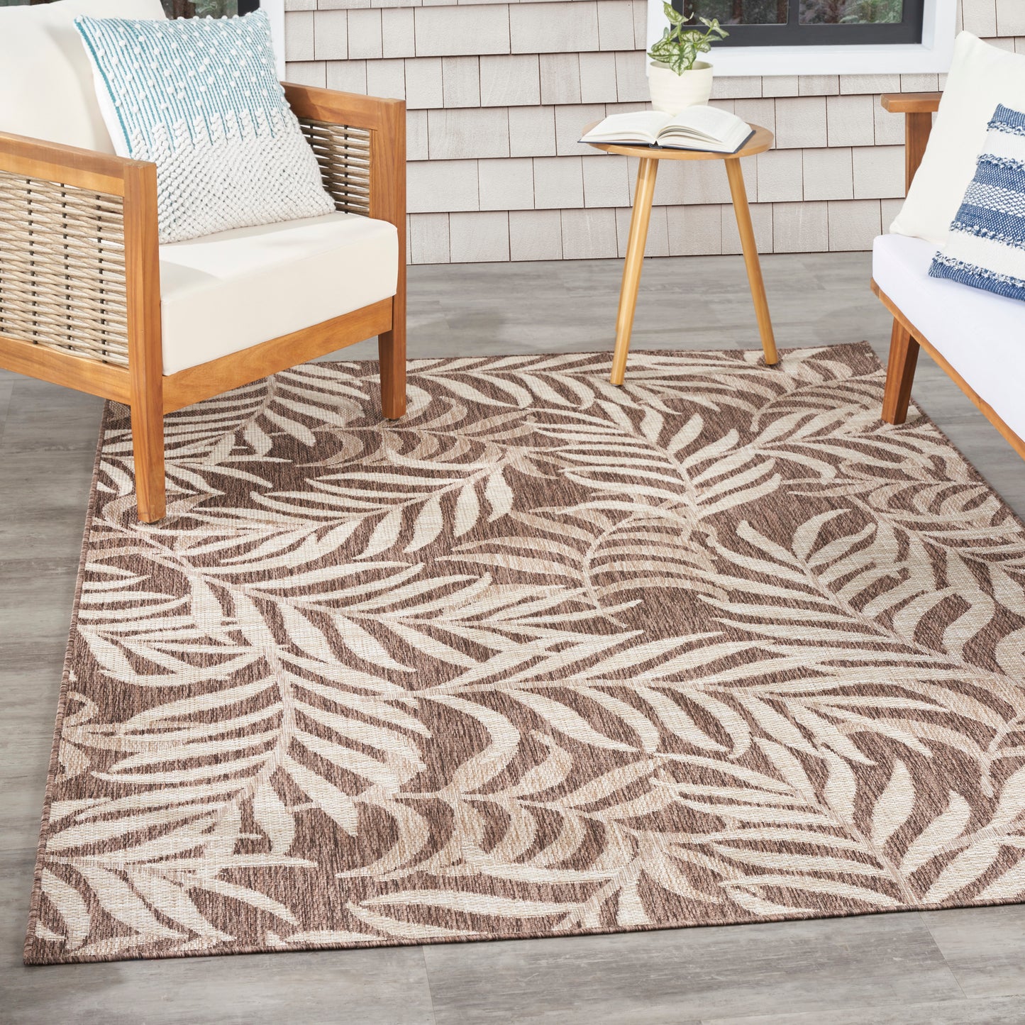 Leaf-patterned brown/beige outdoor rug on gray deck with wooden-framed white cushions and a small side table.