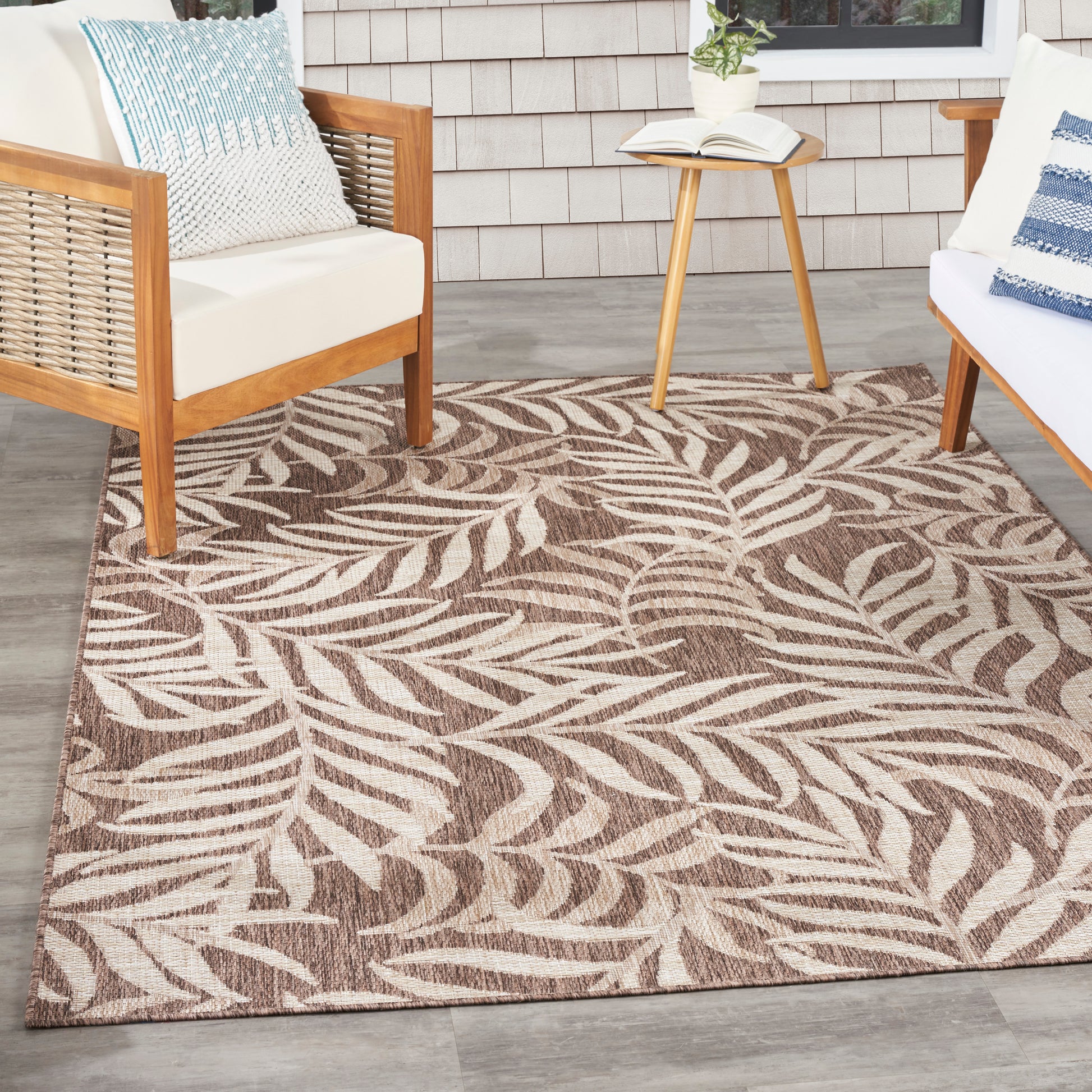 Leaf-patterned brown/beige outdoor rug on gray deck with wooden-framed white cushions and a small side table.