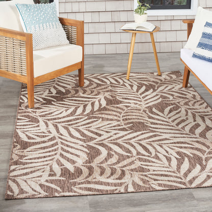 Leaf-patterned brown/beige outdoor rug on gray deck with wooden-framed white cushions and a small side table.