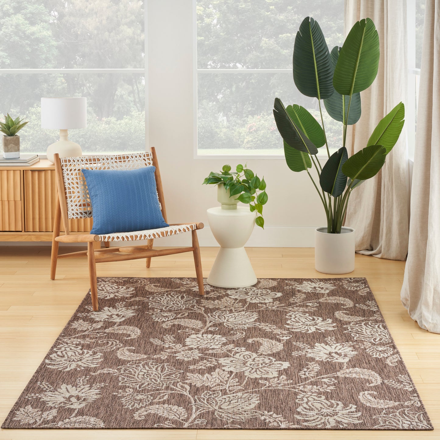 Tropical 5' x 7' rug in a living room: woven brown rug with a beige floral motif, wooden chair with blue cushion and a leafy plant by a window.