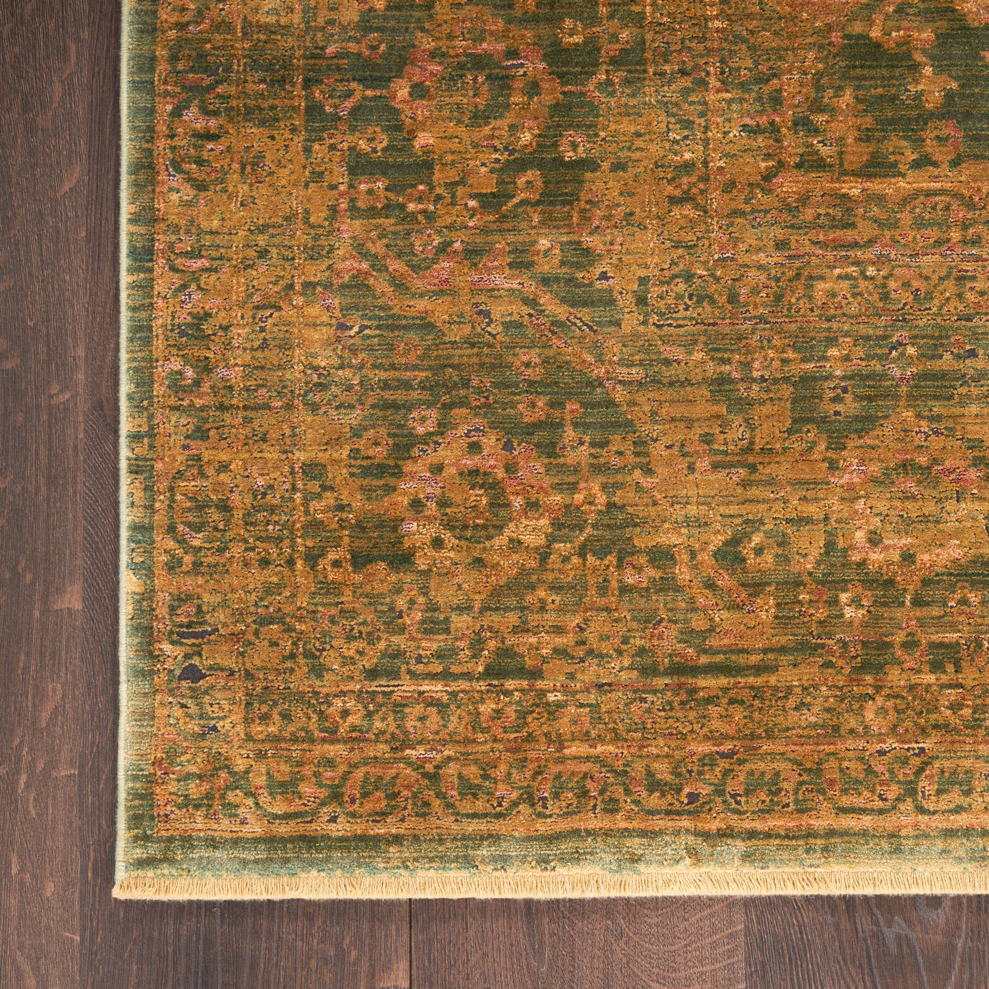 Rectangular oriental rug in olive green, gold, and burnt orange with floral motif and fringed edge on dark wood floor.