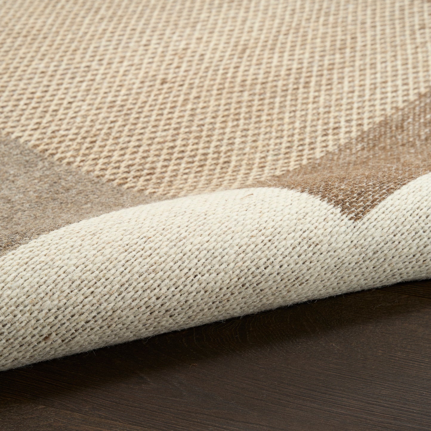 Beige woven area rug with cream and tan geometric pattern, rolled corner on dark wooden floor.