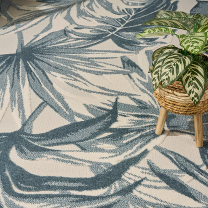 Blue botanical palm-leaf rug with beige background; woven basket plant stand and variegated green-white plant.